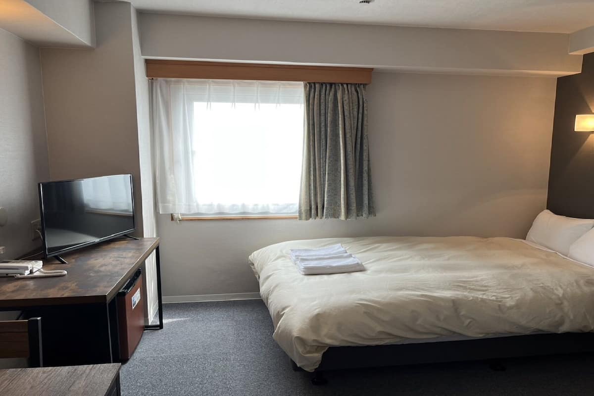 Habitación de hotel con cama, escritorio con televisor, ventana con cortinas y luz en la pared.