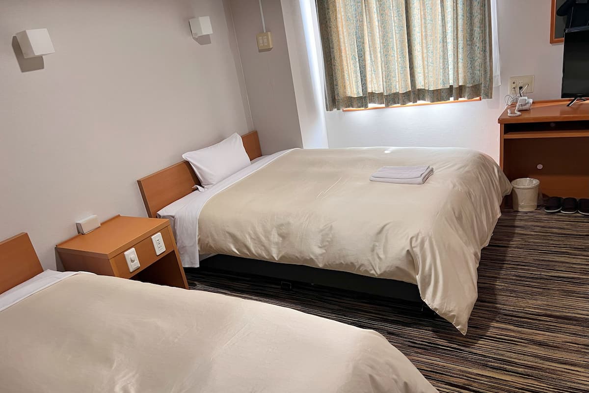 Habitación de hotel con dos camas, una mesita de noche de madera, un escritorio con silla, un cubo de basura y una ventana con cortinas.