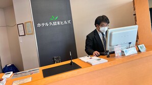 A hotel reception desk with a man working on a computer. There are framed certificates on the wall, a blue bag, and various papers and a small clock on the desk. - Hotel Kurume Hills (Kurume)