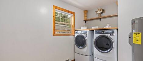 A laundry room with two stacked front-loading washing machines, a window with a wooden frame, and a shelf with laundry supplies.