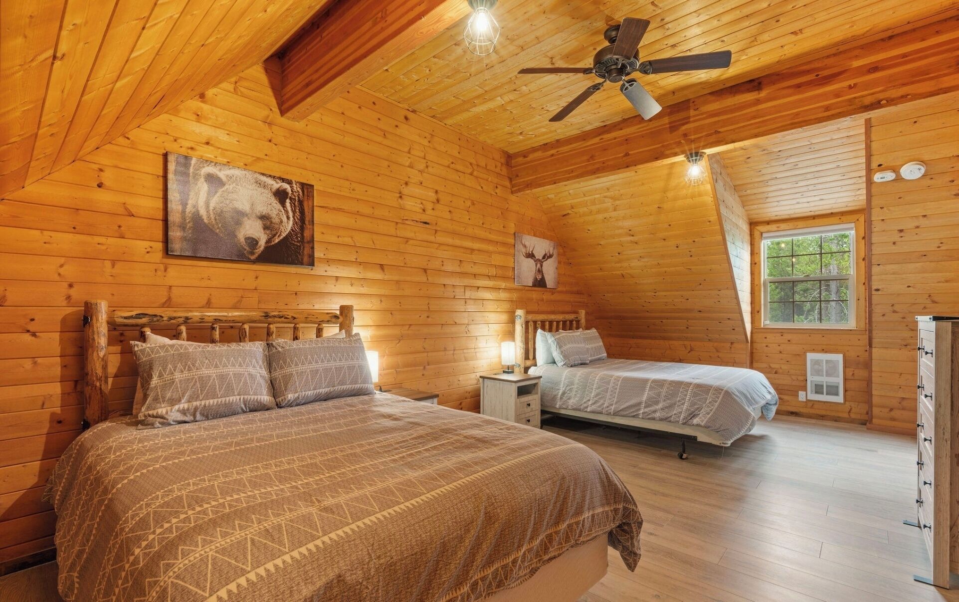 A cozy cabin with two beds, wooden walls, a ceiling fan, and a window with a view of trees.