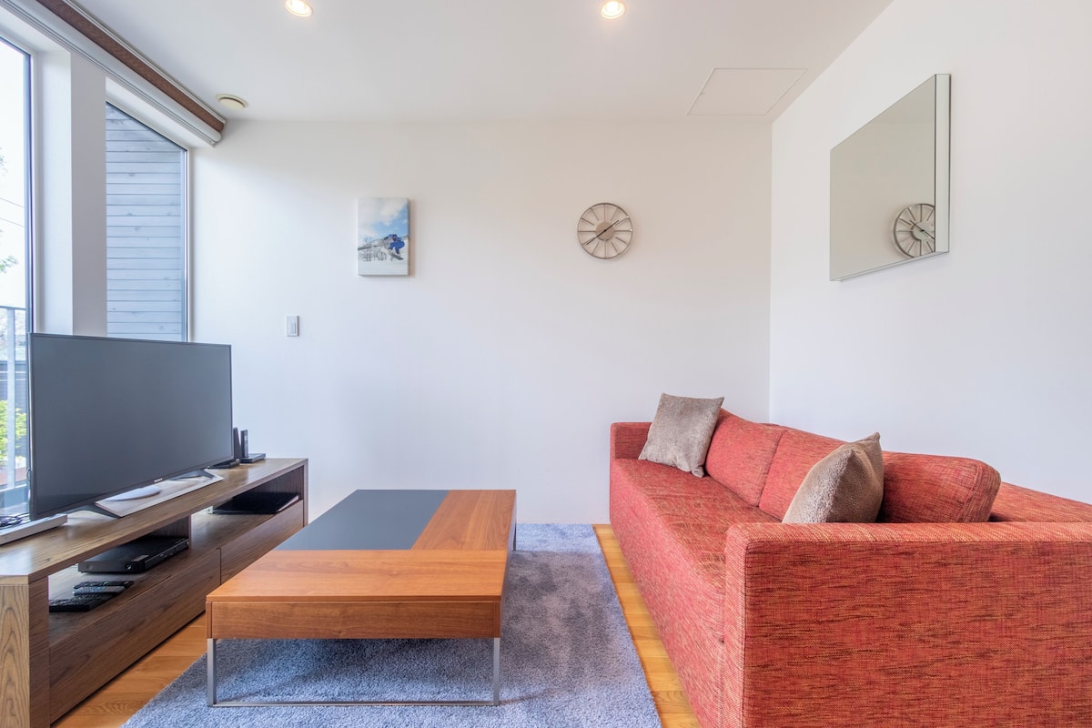 A modern living room with a flat-screen TV on a wooden stand, a wooden coffee table, a red sofa with cushions, and a large window providing natural light.