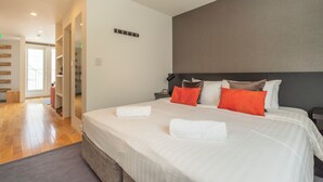 A modern hotel room with a large bed, a bedside table, and a nightstand. There is a door leading to another room and a hallway with wooden flooring. - Hangetsu Niseko (Kutchan)