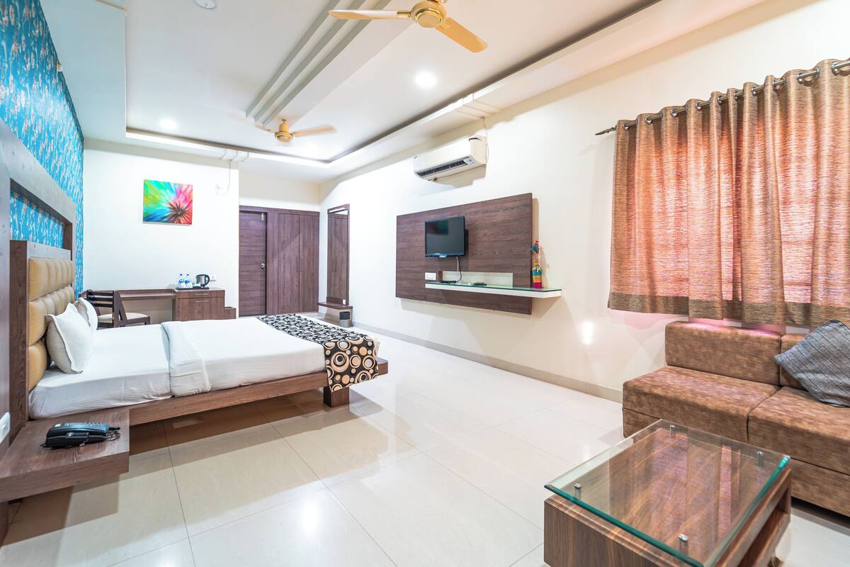 A modern hotel room with a bed, a sofa, a glass coffee table, a TV mounted on the wall, a ceiling fan, and a window with curtains.