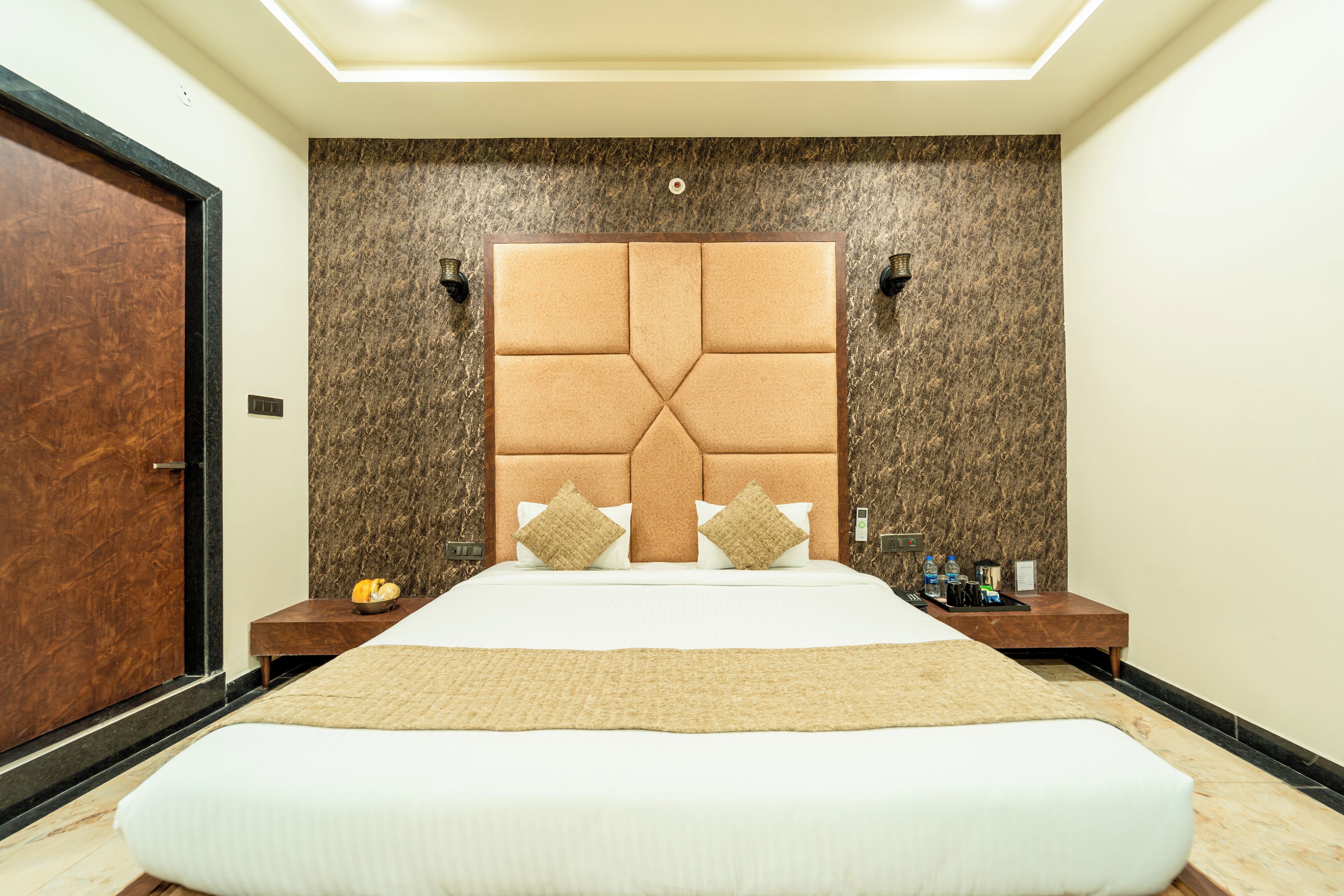 Una habitación de hotel con una cama grande, una puerta de madera, una pared de mármol y una mesita de noche con una lámpara y un atril con artículos de tocador.