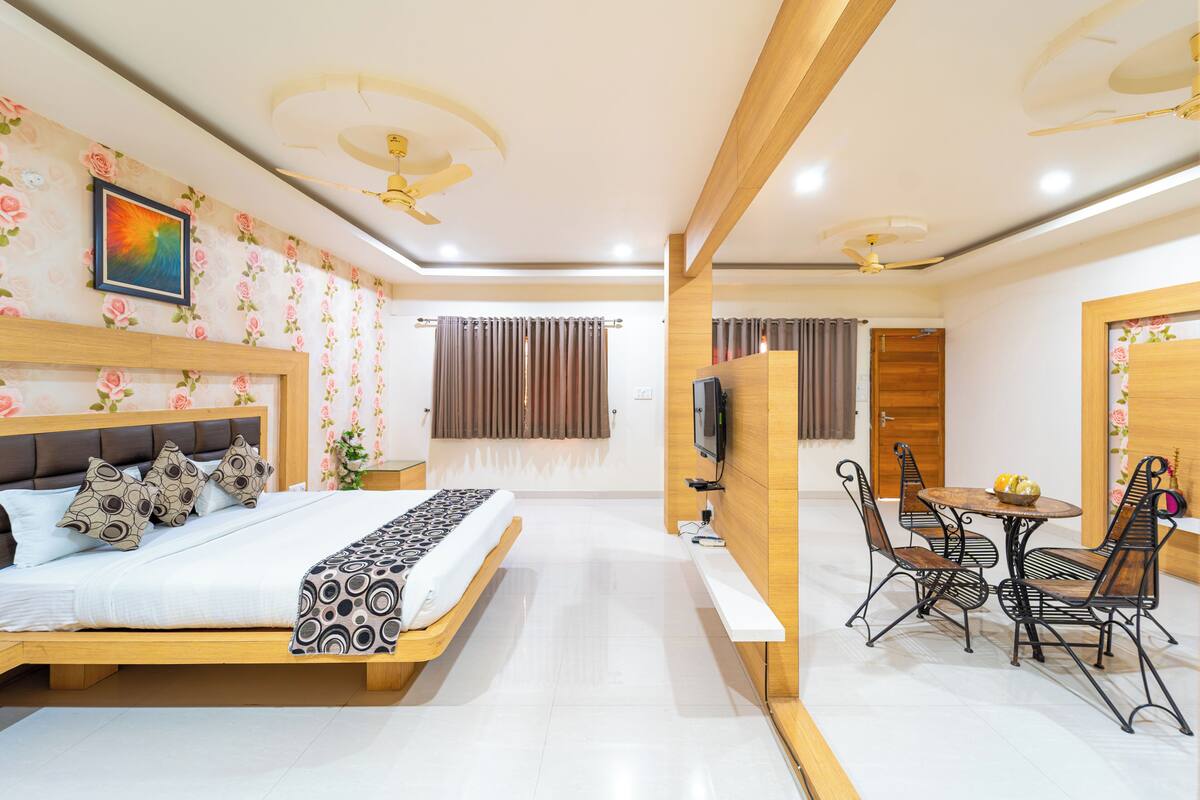 A modern bedroom with a wooden bed, a TV, a dining table with chairs, and a ceiling fan.