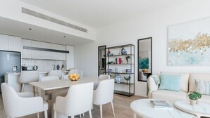A modern, well-lit living area with a dining table, white chairs, a sofa, and a coffee table. There is a bookshelf with decorative items and a painting on the wall.