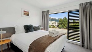 A hotel room with a bed, a bedside table, a lamp, a window with curtains, and a view of the outside. - Ruru Retreat (Wanaka)