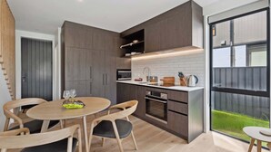 A modern kitchen with dark wood cabinets, a white countertop, and a large window offering a view of a green landscape. - Ruru Retreat (Wanaka)
