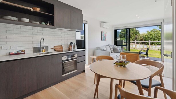 A modern kitchen with dark cabinetry, white subway tiles, and a wooden dining table with chairs. - Ruru Retreat (Wanaka)