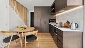 A modern kitchen with wooden cabinets, a white countertop, and a built-in stove. There is a dining area with a round table and chairs. - Ruru Retreat (Wanaka)