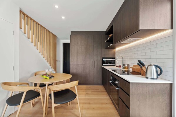 A modern kitchen with wooden cabinets, a white countertop, and a built-in stove. There is a dining area with a round table and chairs. - Ruru Retreat (Wanaka)