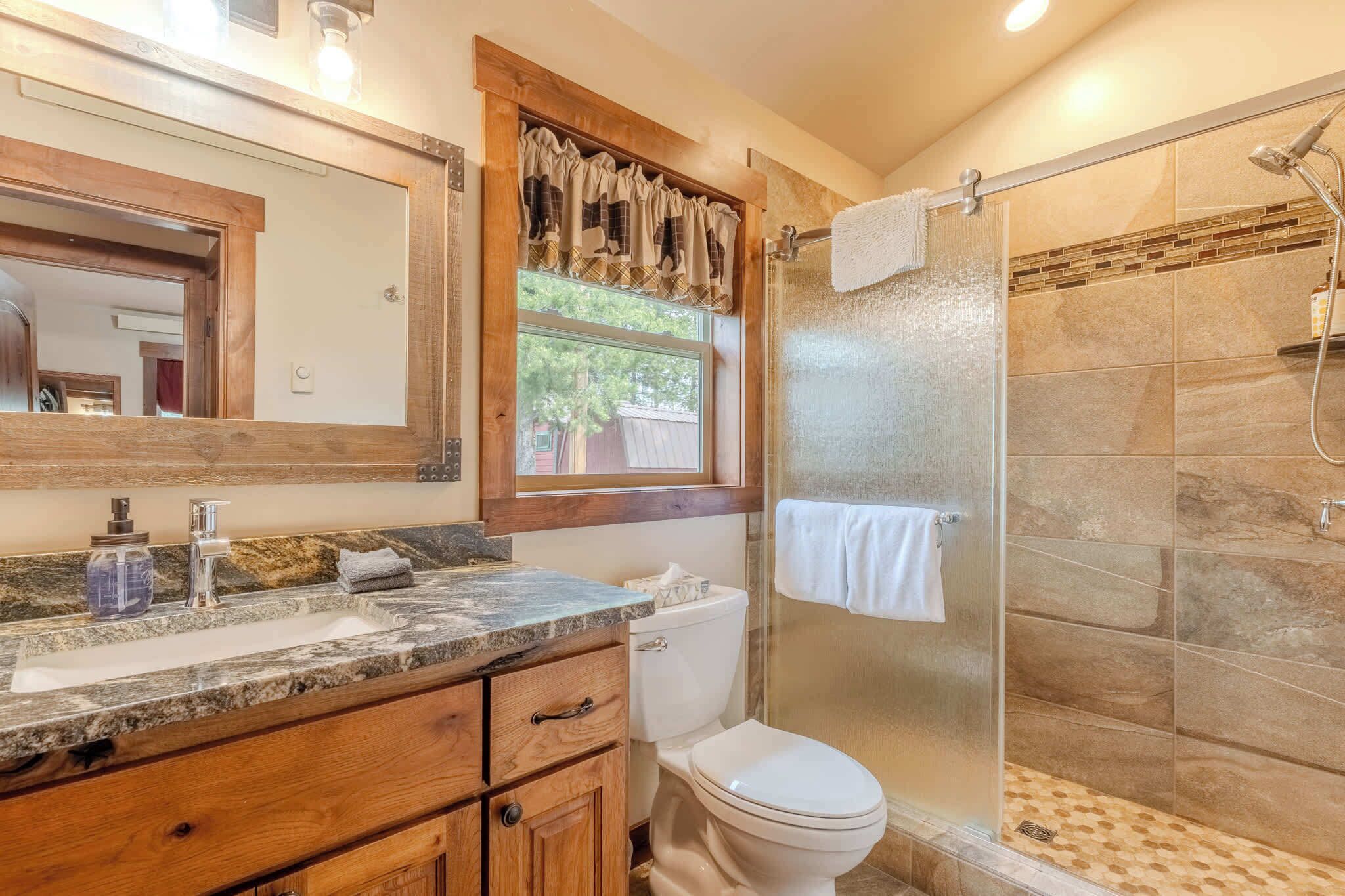 A bathroom with a granite countertop, wooden cabinets, a white toilet, a glass-enclosed shower, and a window with a curtain.