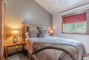 Um quarto com cama, duas luminárias de cabeceira, mesa de cabeceira, janela com valência vermelha e vista para árvores.