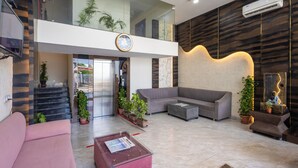 A modern lobby with a grey sofa, a coffee table, potted plants, a staircase, and a clock on the wall. - Hotel Pravasa Stay, Dewas Naka (Indore)
