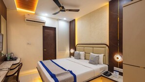 A hotel room with a bed, desk, chair, ceiling fan, air conditioning unit, bedside lamp, and a nightstand with a phone. - Hotel Pravasa Stay, Dewas Naka (Indore)