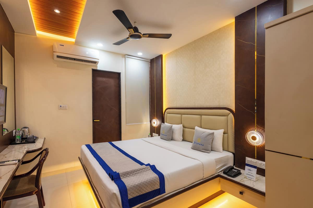A hotel room with a bed, desk, chair, ceiling fan, air conditioning unit, bedside lamp, and a nightstand with a phone.