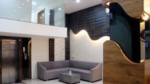 A modern lobby with a grey sectional sofa, a glass coffee table, and a wooden accent wall. - Hotel Pravasa Stay, Dewas Naka (Indore)