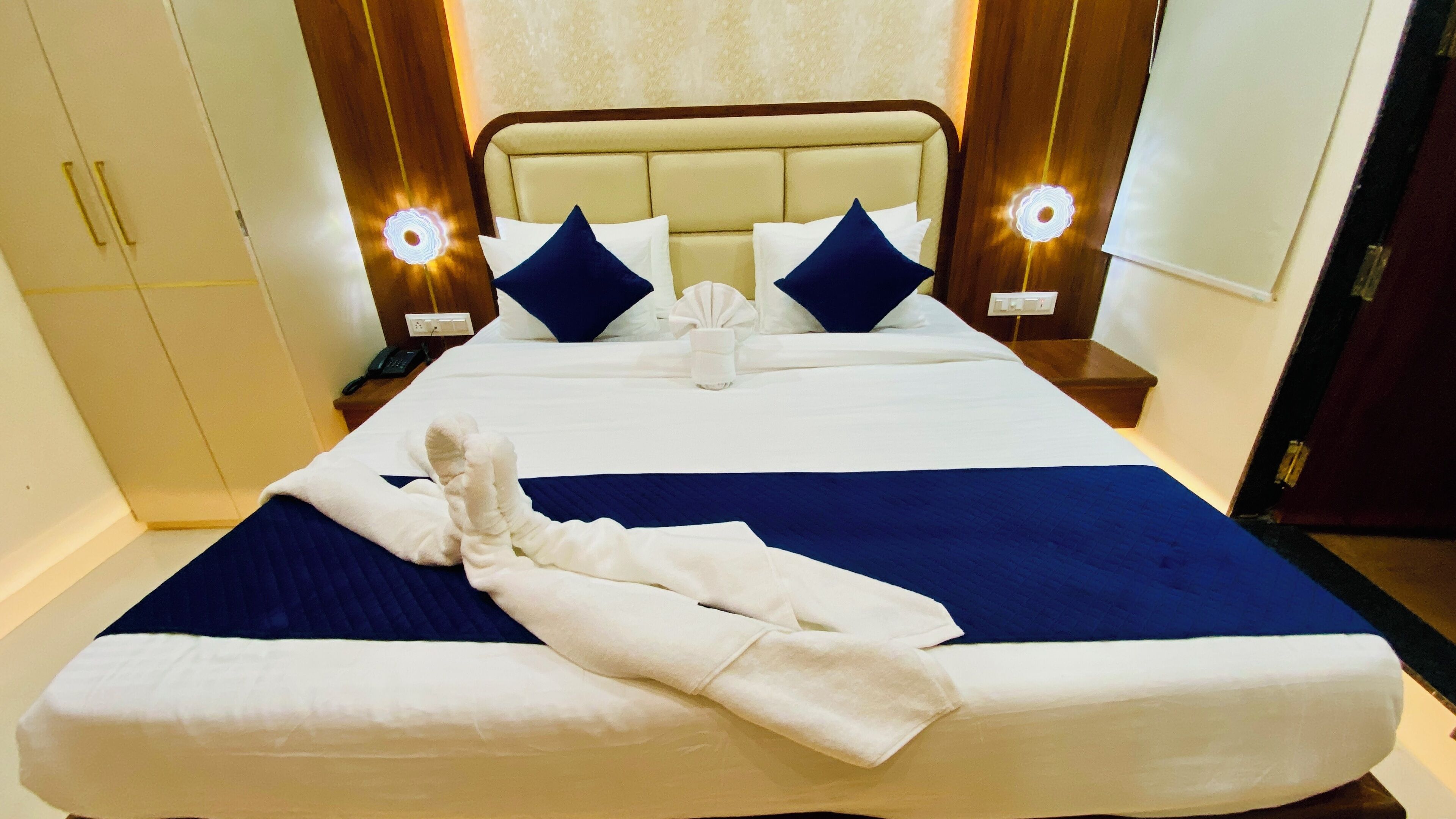 Un letto rifatto con cura, coperto da una trapunta blu e bianca, un asciugamano bianco ripiegato e due cuscini blu. Sono presenti due abat-jour con un design luminoso circolare, un comodino in legno con un telefono e un armadio in legno.
