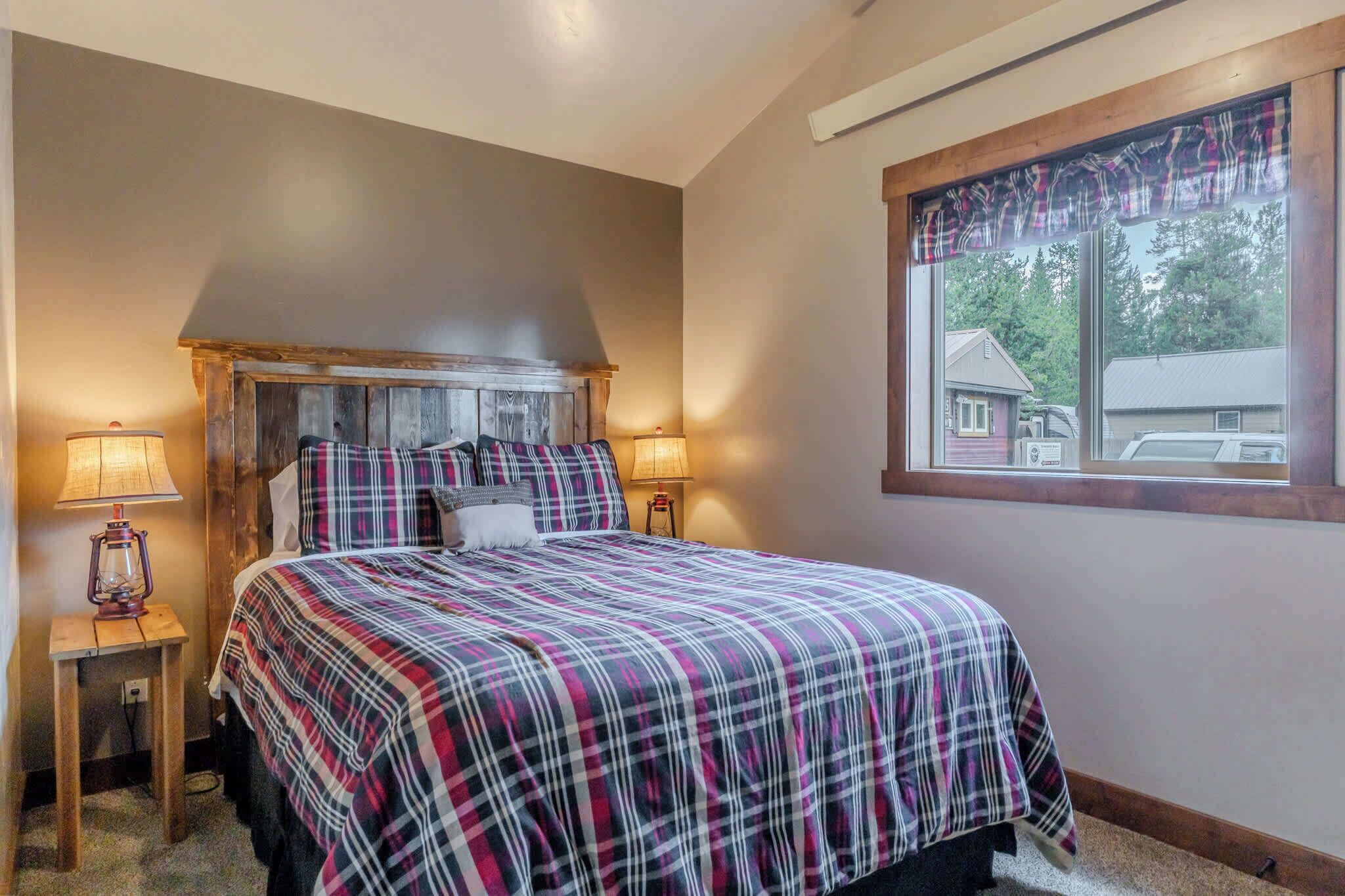 A bedroom with a bed, two bedside lamps, a window with a view, and a plaid bedspread.