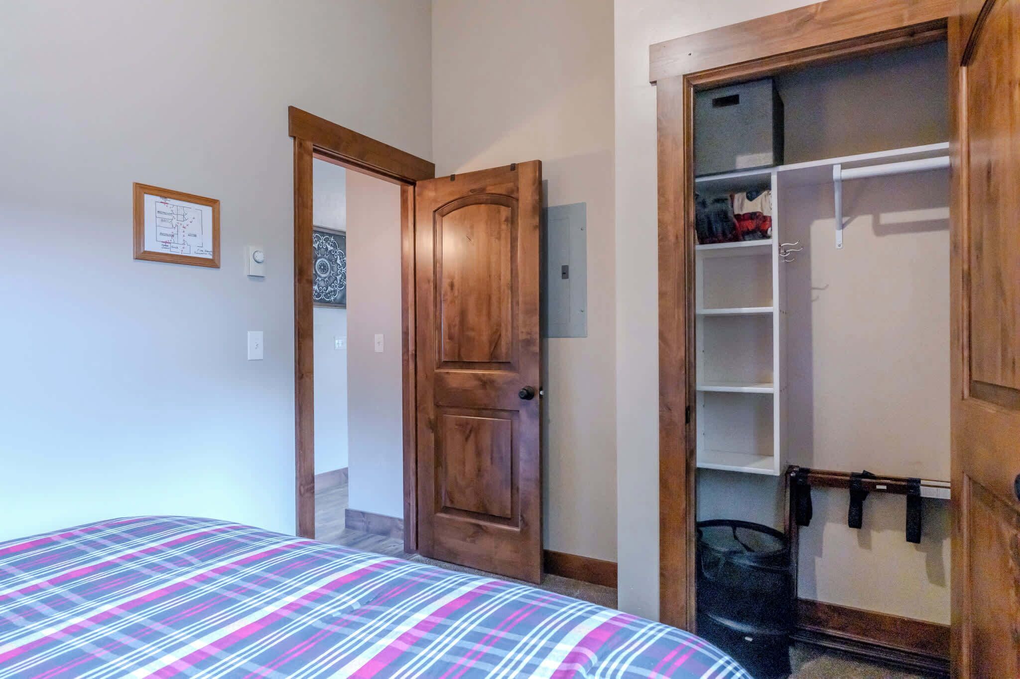 A bedroom with a bed, a wooden door, a closet with hanging items, and a wall-mounted light switch.