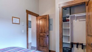 A bedroom with a bed, a wooden door, a closet with hanging items, and a wall-mounted light switch.