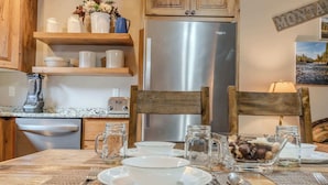A kitchen with wooden cabinets, a stainless steel refrigerator, a blender, and a dining table set with plates, cups, and a bowl of nuts.