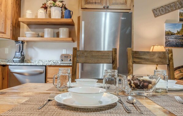 A kitchen with wooden cabinets, a stainless steel refrigerator, a blender, and a dining table set with plates, cups, and a bowl of nuts.