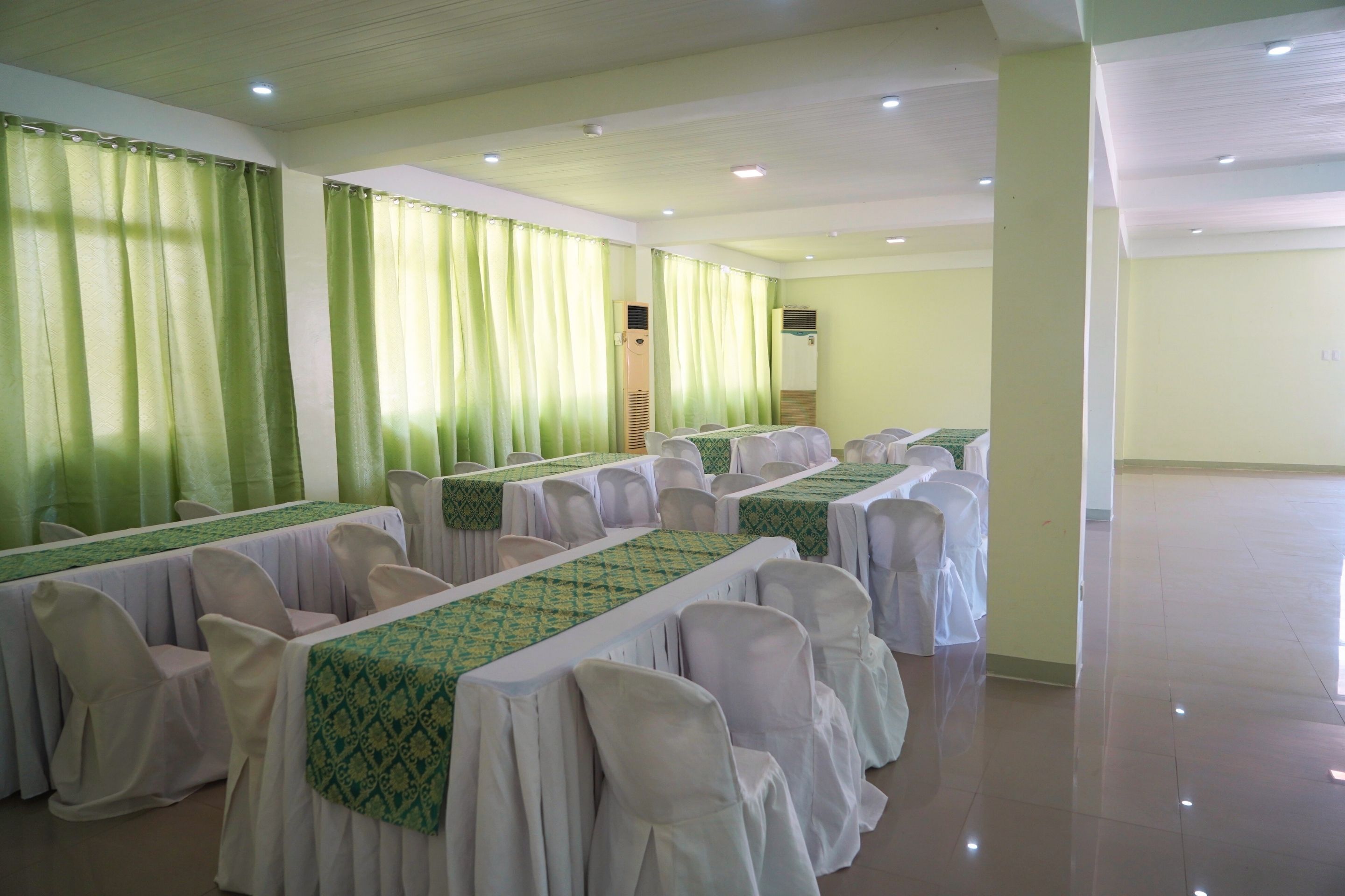 Un salón de conferencias con mesas dispuestas para una reunión, sillas con fundas blancas y cortinas verdes.
