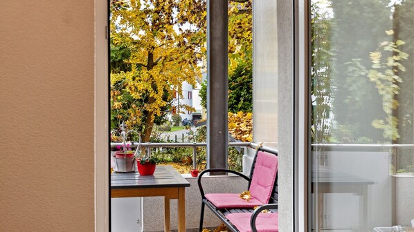 Un balcón con una mesa y sillas, plantas en macetas y vistas a árboles y edificios.