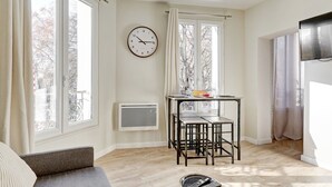 A room with a large window, a wall clock, a table with chairs, and a radiator. - Mark 22 - Large studio Canal Saint-Martin views (Paris)
