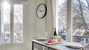 A dining area with a table set for two, a clock on the wall, and a view of the street outside. - Mark 22 - Large studio Canal Saint-Martin views (Paris)