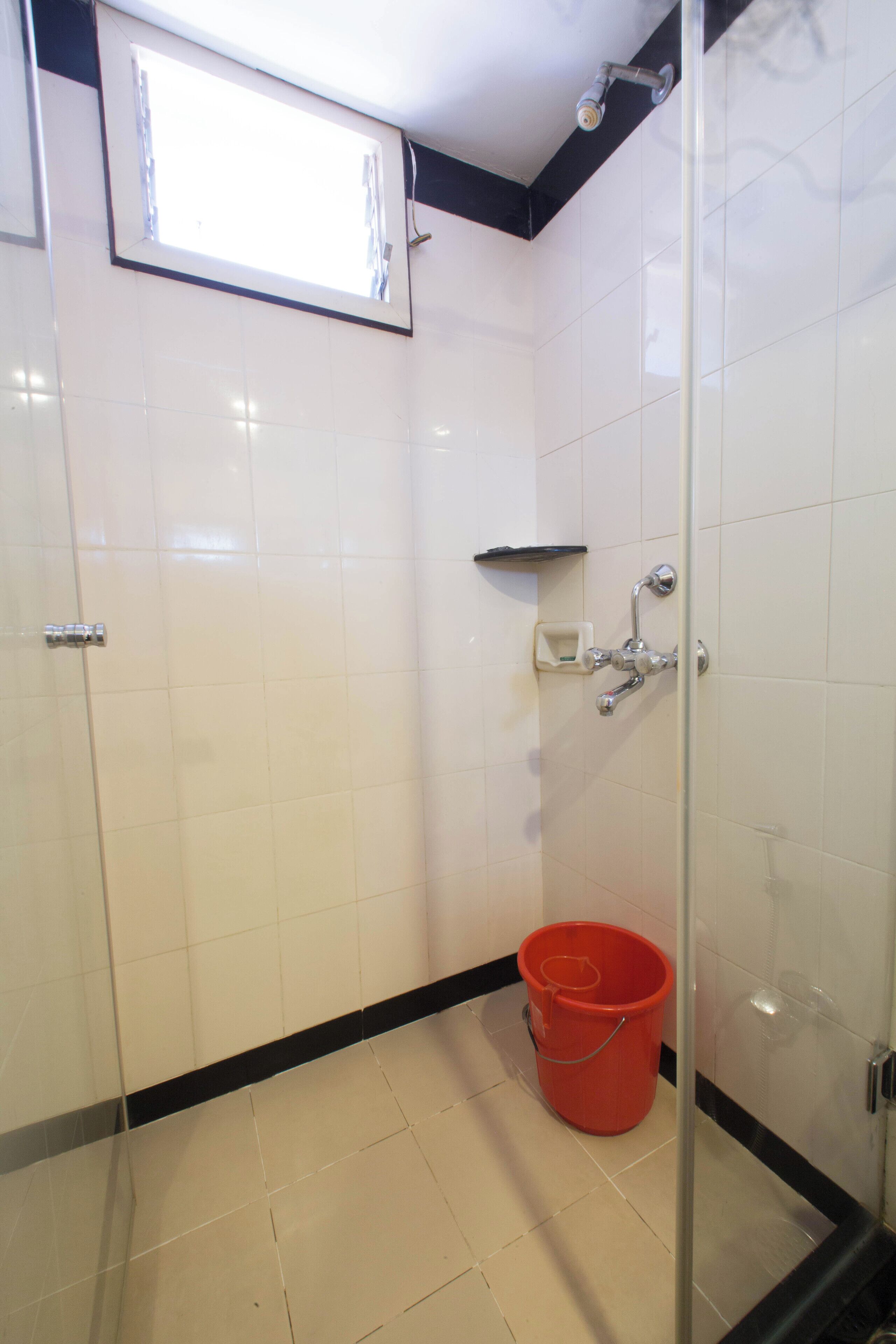 Un baño con azulejos blancos, cabina de ducha de vidrio, un cubo rojo y una pequeña estantería.