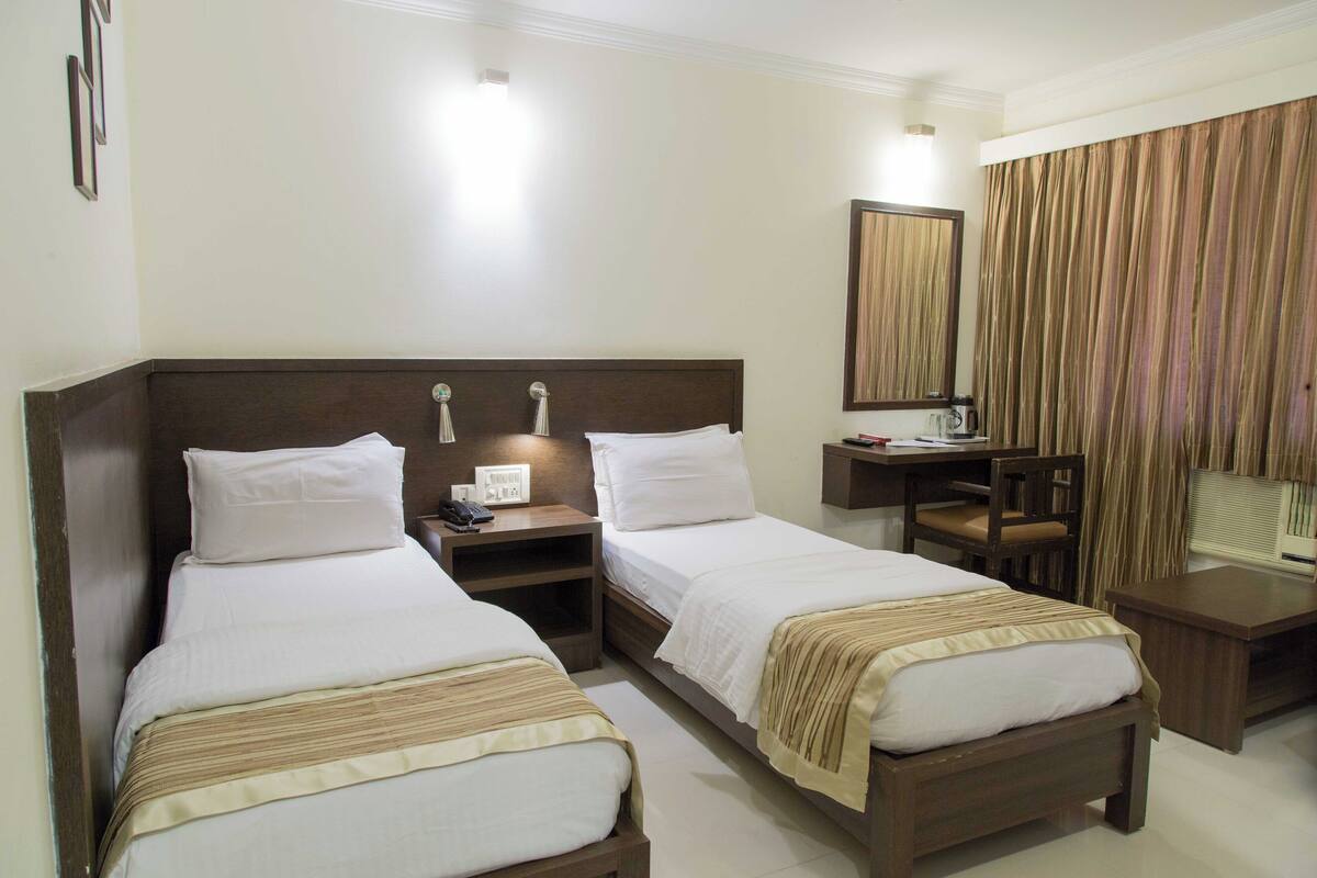 A hotel room with two single beds, a desk, a chair, a mirror, and a window with curtains.