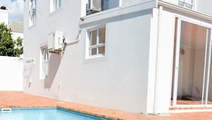 A swimming pool adjacent to a white building with multiple windows and an air conditioning unit. - Portmarnock House (Cape Town)