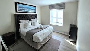 A bedroom with a large bed, a window with blinds, a framed picture on the wall, and a small bedside table. - Portmarnock House (Cape Town)