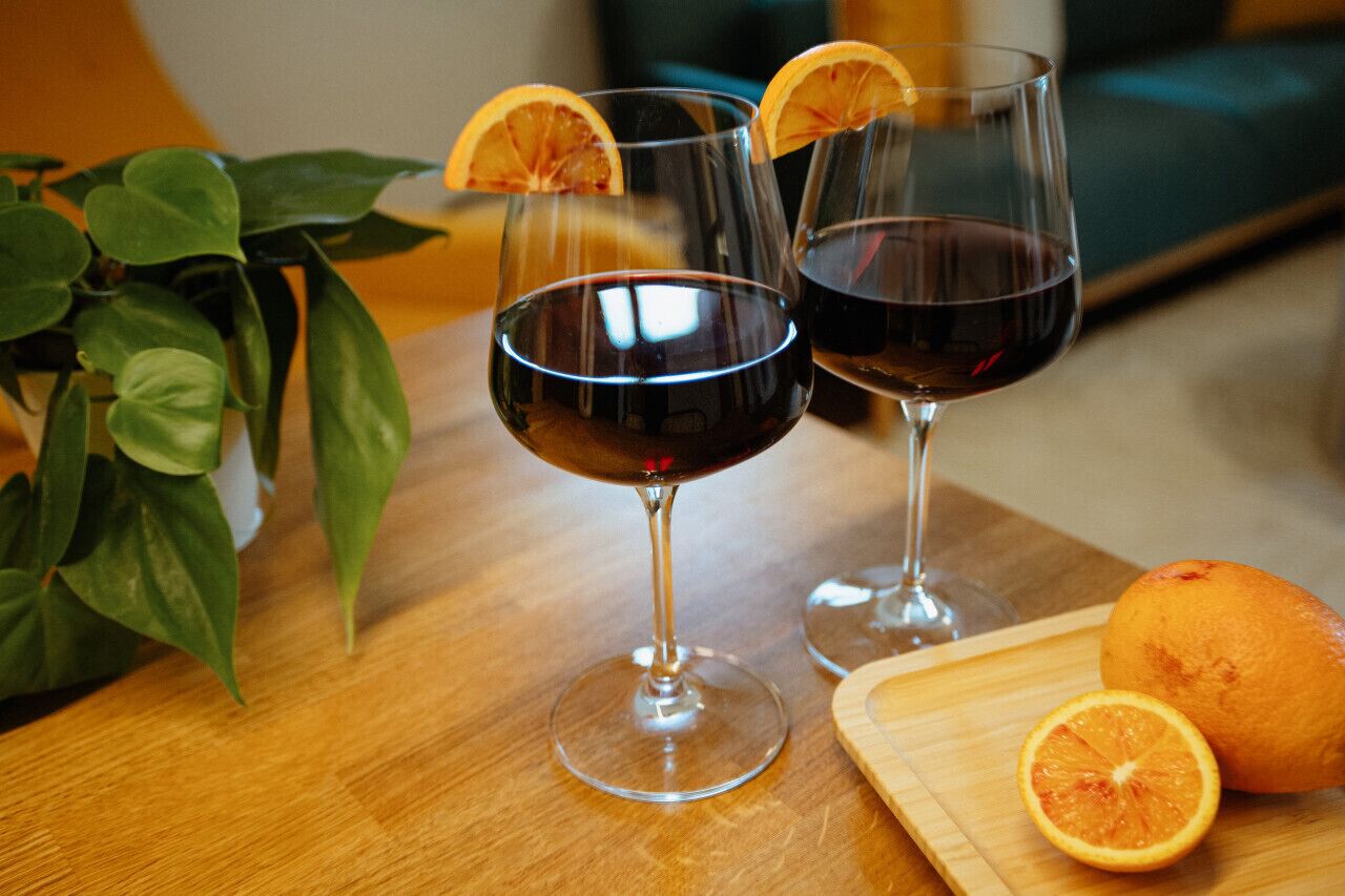 Two wine glasses with a dark liquid, a potted plant, and a wooden cutting board with an orange and its slice.
