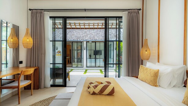 A modern hotel room with a large bed, a wooden desk, and a view of the garden through glass doors. - Milla Villa by Betterplace (Ubud)