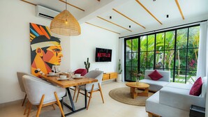 A modern living room with a dining area, a sofa, a TV, and a large window with a view of greenery. - Milla Villa by Betterplace (Ubud)