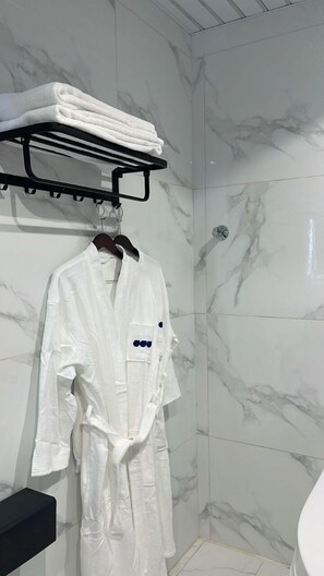 A bathroom with a marble shower wall, a white robe hanging on a rack, and a towel shelf above it. - Qimam Park 5 Abha (Abha)