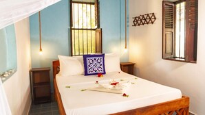 A neatly made bed with a white bedspread, a blue decorative pillow, and a wooden headboard. There is a small wooden nightstand beside the bed, and a wall-mounted shelf above it. - Crystal Blue - Comfort Double Room Sea View with AC (Jambiani)
