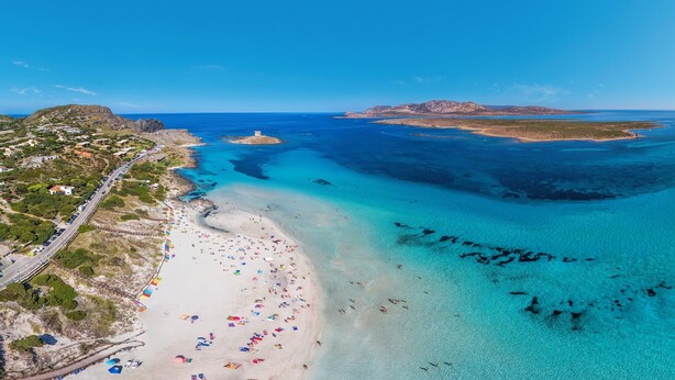 An aerial view of a coastal area with a beach, clear blue waters, and a road leading to a settlement.