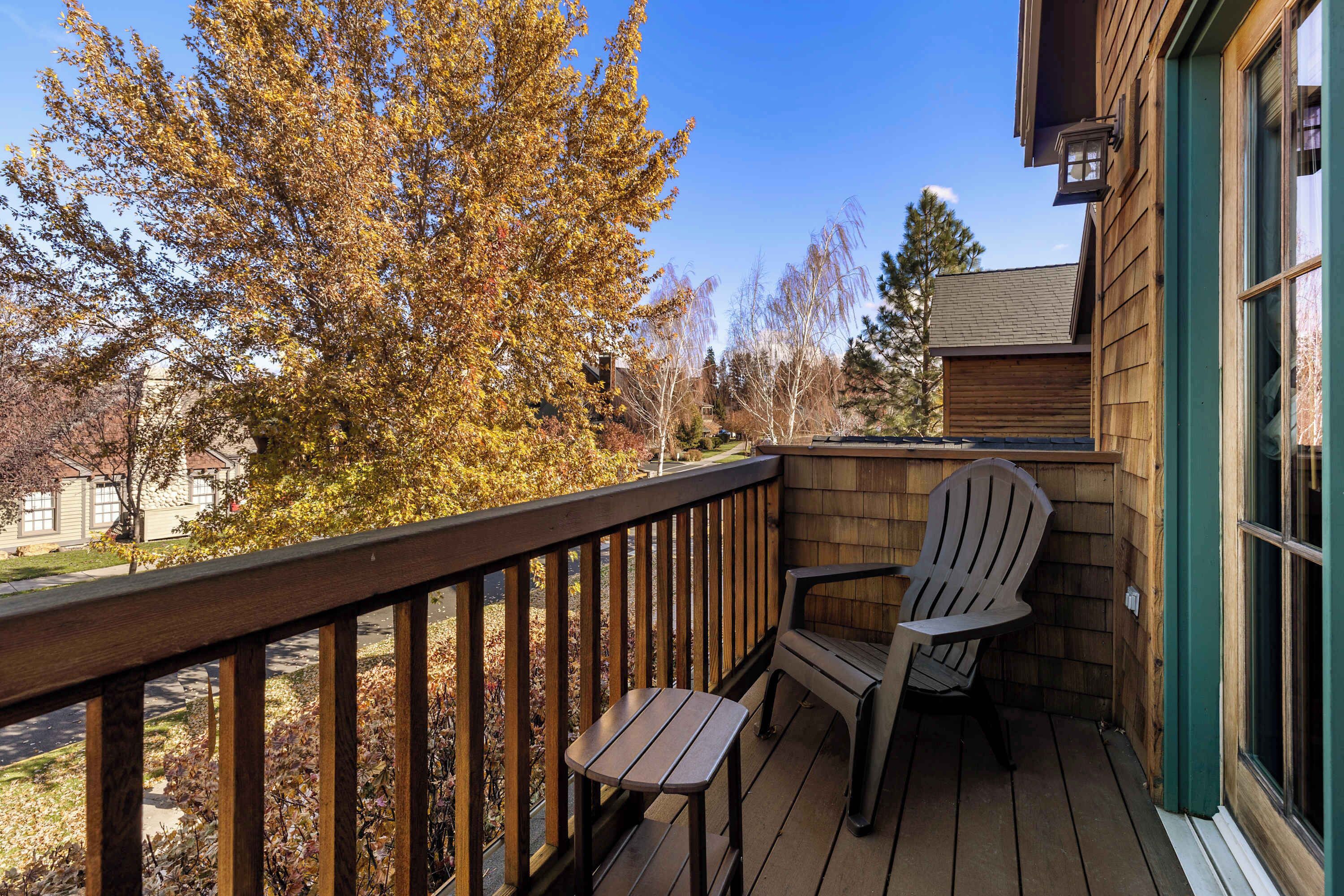 A wooden deck with a chair and table, overlooking a street and autumn trees.