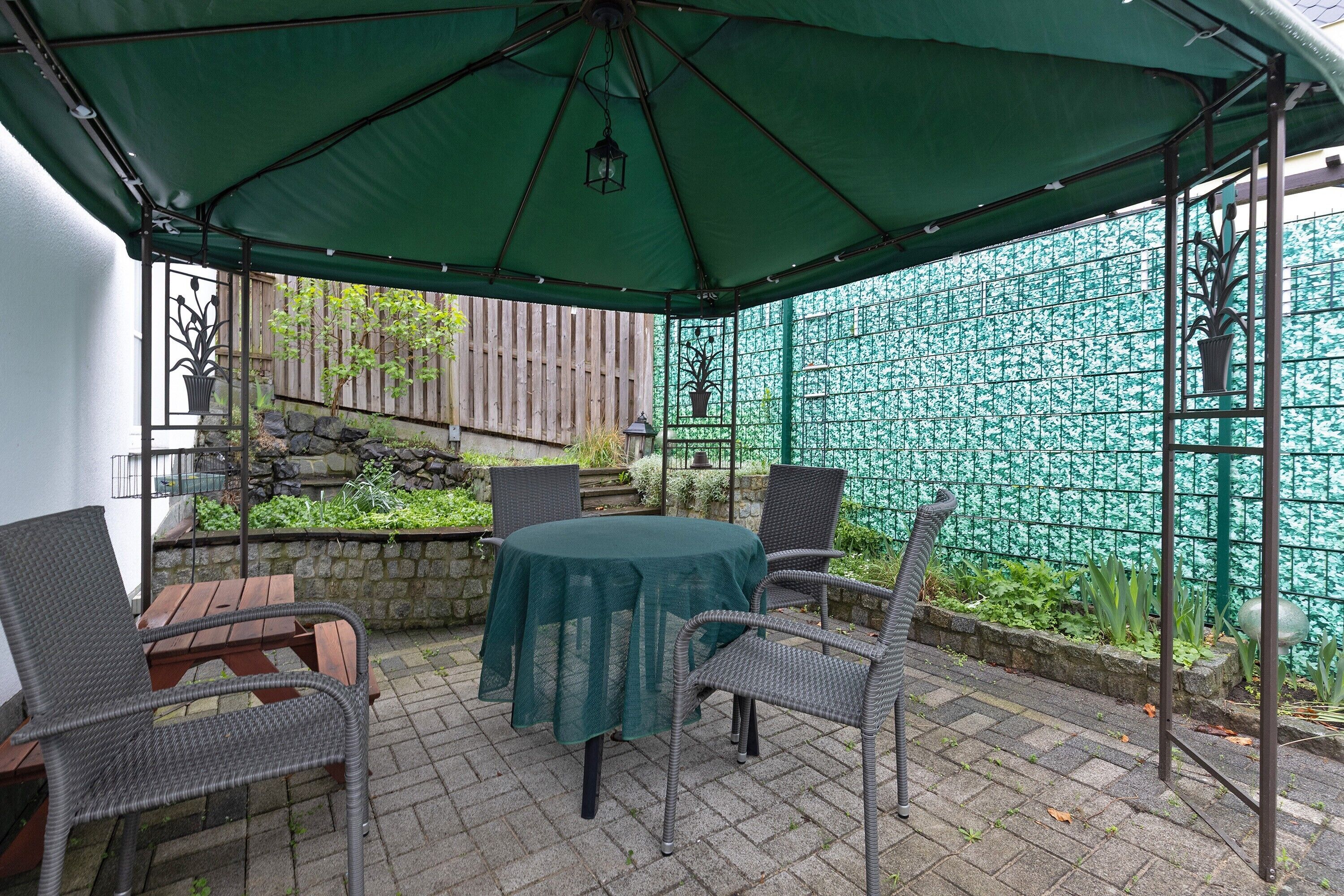 En terrasse med et bord og stole under et grønt markise, omgivet af en stenvæg og et metalhegn.