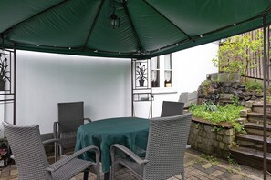A covered patio with wicker furniture and a table, a stone wall with plants, and a view of a building with windows.
