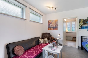 Ein modernes Wohnzimmer mit einem schwarzen Ledersofa, einem kleinen weißen Tisch mit einer Mäusefigur, einer roten, gemusterten Decke und einem Gemälde an der Wand.