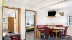 A dining area with a wooden table and red chairs, a sofa with a red pillow, and a television mounted on the wall.