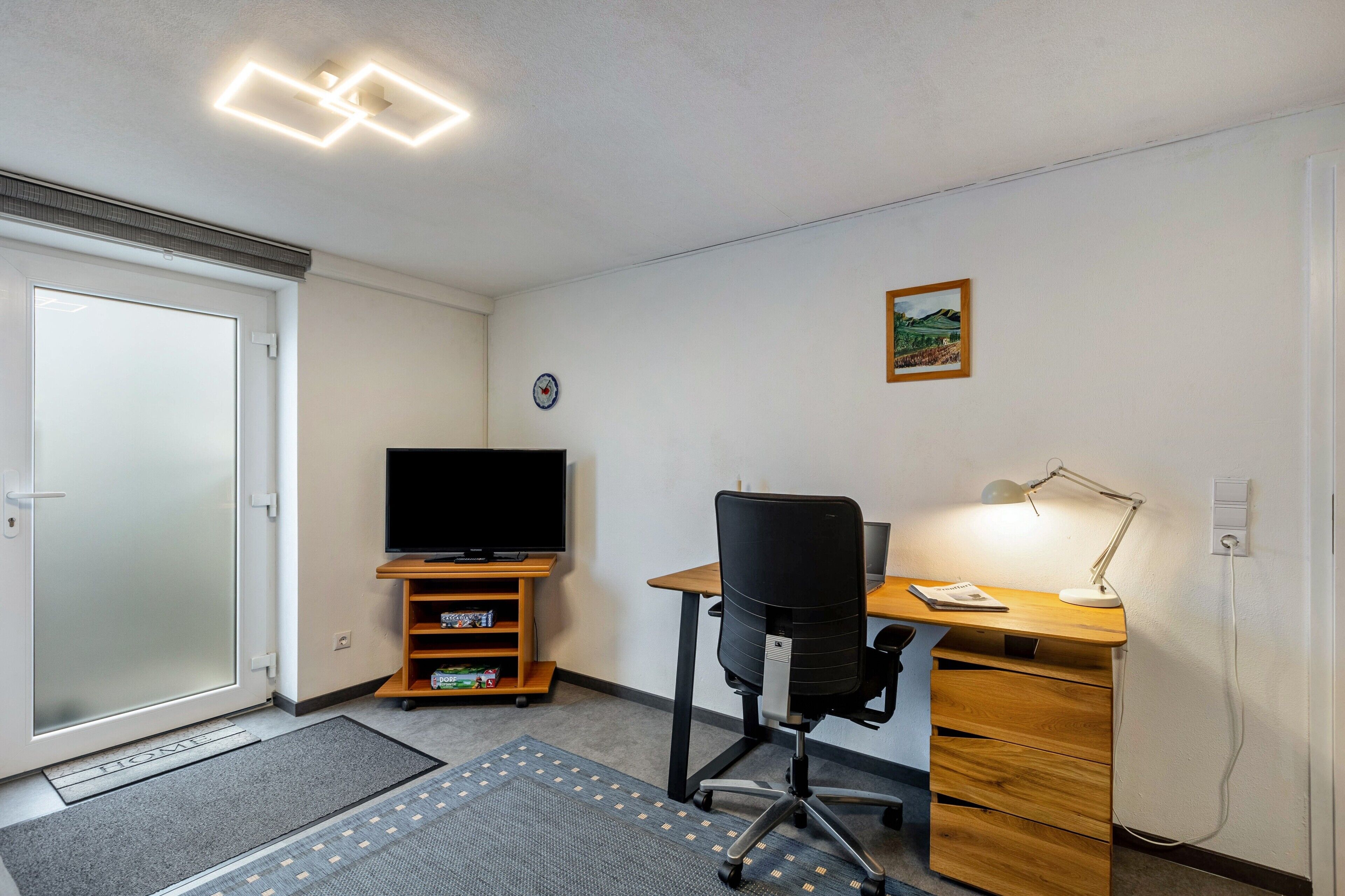 Une pièce comprenant un bureau, une chaise, un téléviseur et une petite étagère avec quelques objets.