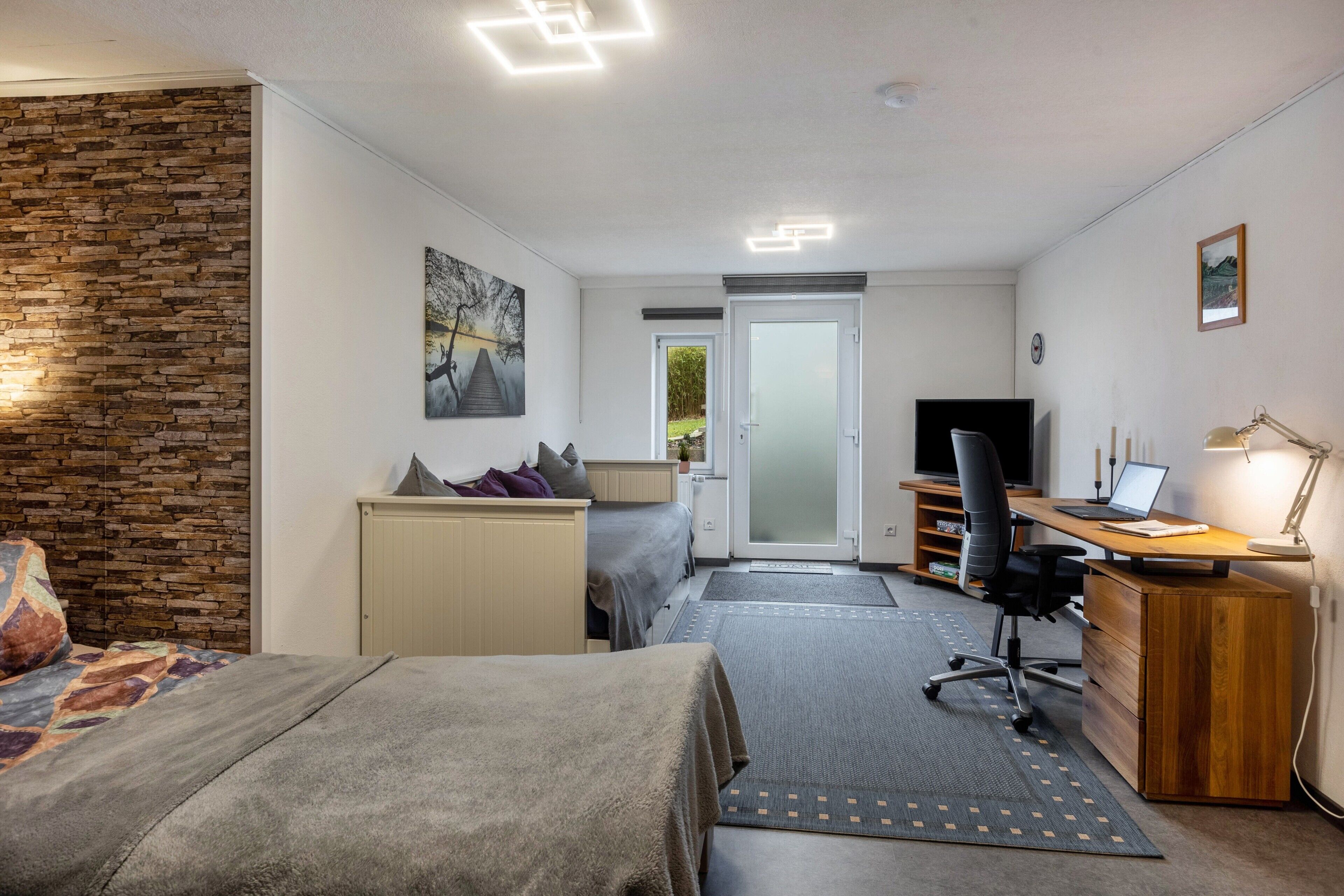 A bedroom with a bed, a desk, a chair, a television, and a door leading to a balcony.