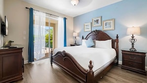 A bedroom with a large bed, wooden headboard, two bedside tables with lamps, a desk with a chair, and a sliding glass door leading to a balcony.